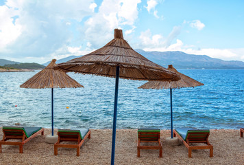 View of empty beach – sun bads and umbrellas near sea water, pebbles and golden sand, mountains on the horizon and clouds on the sky. Landscape of abandoned summer resort.