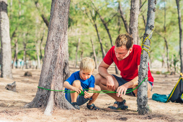 Naklejka na ściany i meble Camping people outdoor lifestyle tourists in summer forest near lazur sea. Blond boy son with father study survival techniques, practice methods of tying rope knots. Natural children education