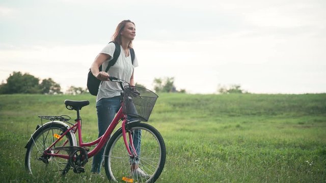 Cinematic shot of a young woman walking on a green field with a bicycle on sunset time.