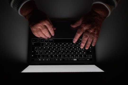 Elderly woman's hands on a computer keyboard in the dark, light from the screen. The older generation is searching for information on working and studying freelance on the Internet using a laptop