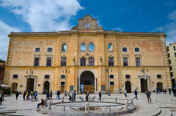 Matera, Italy - May 6, 2018: People walking near Cinema Comunale Palazzo dell'Annunziata palace with clock on facade and fountain on Piazza Vittorio Veneto square blue sky background