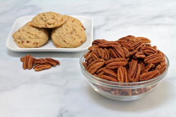 Half pecans in a glass  bowl with pecan cookies in the background.
