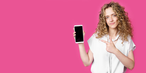 Look at this cell phone. Contented happy woman, pointing her index finger at a blank screen, shows a modern device. Isolated on a pink background.