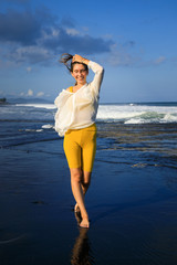 Beautiful young woman walking on black sand beach. Caucasian woman wearing yellow sportswear and white blouse. Happiness and freedom. Travel lifestyle. Copy space.