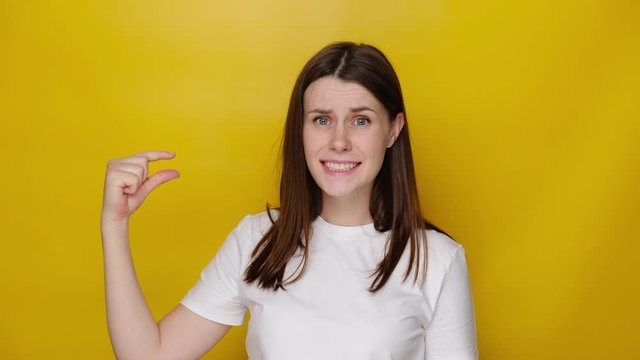 Brunette young woman isolated over yellow studio background shows very tiny object, shapes something small, dressed in white t-shirt, female looks in puzzlement, need some more. I need little bit