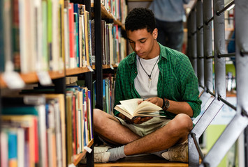 Young male student study in the library reading book while sitting near bookshelf.