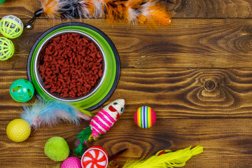 Set of toys for cat and bowl with dry pet food on wooden background. Top view, copy space. Pet care concept