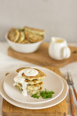 Zucchini fritters with herbs on a light plate and a wooden board, in the background a gravy boat with sour cream and a bowl of fritters