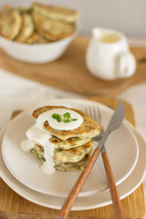 Zucchini pancakes with herbs on a light plate and a wooden board with cutlery, in the background a gravy boat with sour cream and a bowl of pancakes close-up