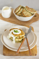 Zucchini pancakes with herbs on a light plate and a wooden board with cutlery, in the background a gravy boat with sour cream and a bowl of pancakes