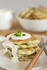 Zucchini pancakes with herbs on a light plate and a wooden board with cutlery, in the background a gravy boat with sour cream and a bowl of pancakes close-up