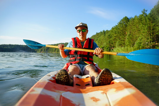 the guy is sailing on a red kayak, wearing a life jacket with glasses and a cap. Holds a paddle in his hand