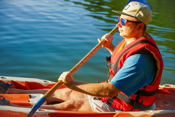 the guy is sailing on a red kayak, wearing a life jacket with glasses and a cap. Holds a paddle in...