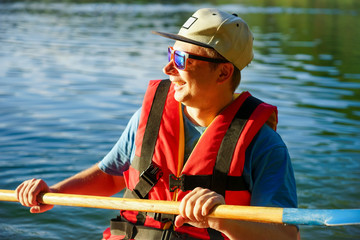 Fototapeta premium the guy is sailing on a red kayak, wearing a life jacket with glasses and a cap. Holds a paddle in his hand