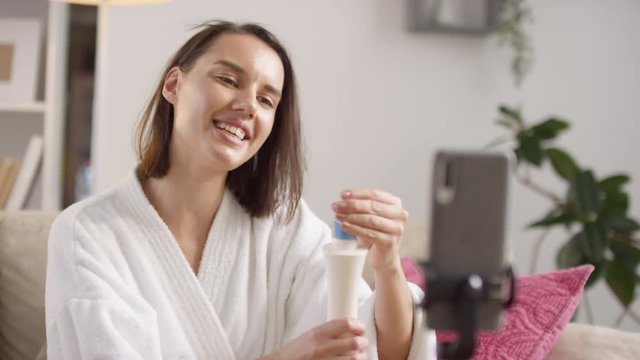 Beautiful young woman wearing white bath robe reviewing protective hair primer spray on smartphone camera