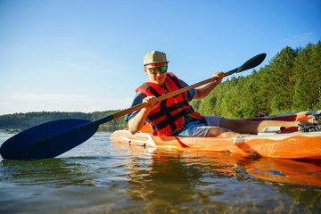 the guy is sailing on a red kayak, wearing a life jacket with glasses and a cap. Holds a paddle in...