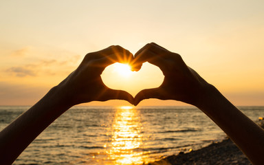 Human hands show the shape of a heart at sunset against the background of the sea. Sun rays at sunset.