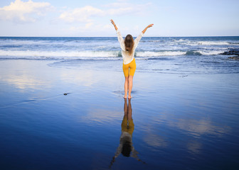 Beautiful young woman standing on black sand beach, raising arms up. Caucasian woman wearing yellow sportswear and white blouse. View from back. Travel lifestyle. Copy space..