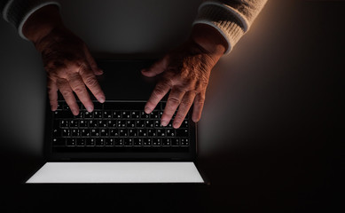 Elderly woman's hands on a computer keyboard in the dark, light from the screen. The older...