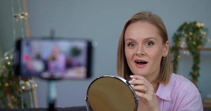 Attractive woman talking and applying mascara while recording video for beauty vlog on smartphone. Cheerful girl vloger sitting in front of mirror while testing new cosmetic.