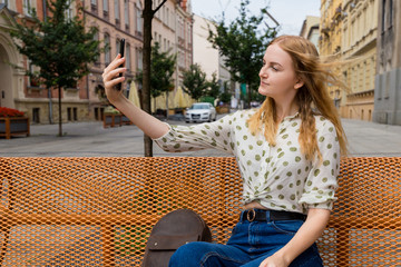 Beautiful young woman with blonde hair taking selfie at the city street background. Girl sitting on a bench in a sunny day. Active life concept