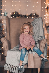 Cute funny baby girl 3-4 year old wearing denim pants and knitted sweater sitting in chair over Christmas glowing lights in room closeup. Winter holiday season. Xmas.