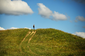 Fototapeta premium A girl with a backpack stands with her back on a hill in the distance among the green grass and flowers.