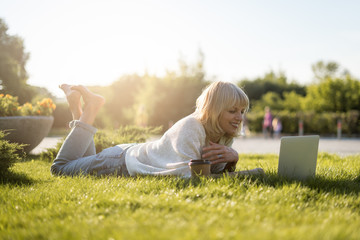 Adult Woman looks at the laptop, lying on the grass outside in park. Happy and smiling senior Working and drinking coffee. Using computer. Distance learning online education and work.