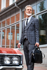 Fototapeta na wymiar elegant senior man with gray hair and beard walks with bag outdoors, next to red luxurious car in the street