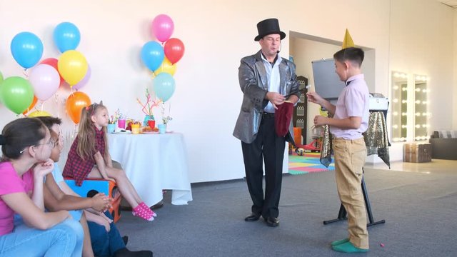 performer in silver jacket and top hat shows magic tricks with little boy help in decorated room at birthday party slow motion