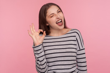 I am okay, approval. Portrait of satisfied happy young woman in striped sweatshirt doing ok gesture and winking to camera, agree with suggestion. indoor studio shot isolated on pink background