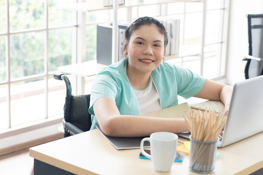 Young Disabled Woman Sit On Wheelchair Using Computer Laptop For Work At Office Workplace, Look At The Camera, Disabled People Concept