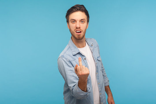 Portrait of aggressive vulgar man in worker denim shirt showing middle finger impolite gesture, demonstrating hate and disrespect, rebellious mood. indoor studio shot isolated on blue background