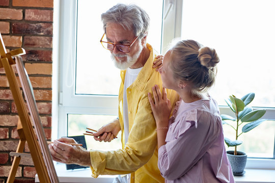 senior man, mature grandfather and his little granddaughter drawing, painting together at home, family time