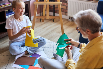 graddaughter and grandfather work with creativity, cutiting the cardboard paper, pleasant nice little girl sitting on the floor with her grandfather