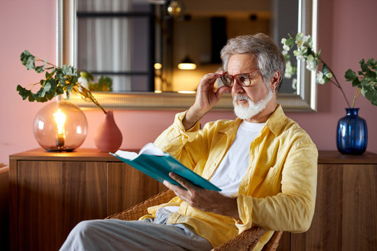 intelligent mature man read book at home, enjoy free time alone, sit on chair, wearing eyeglasses for better reading