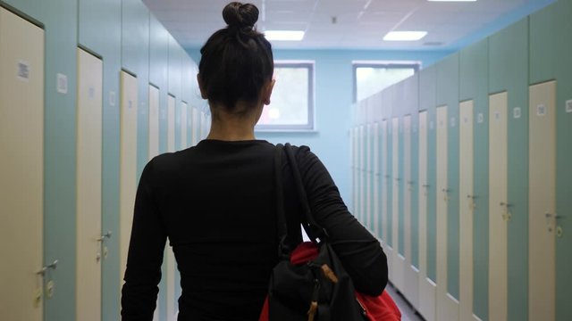 slim girl with bun hairstyle in black clothes walks carrying sports bag along empty brightly lit gym dressing room slow motion backside view
