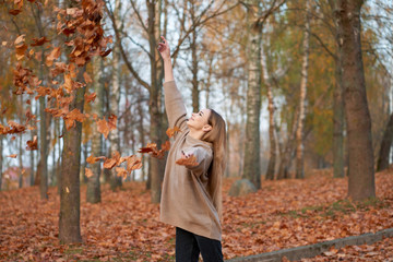 Enjoy autumntime. Beautiful stylish blonde confident girl  wearing sweater, black jeans and leather black boots tossing autumn leaves in autumn park.