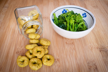 Homemade rapini flavour taralli crackers poured from a glass jar next to a bowl full of boiled rapini. Traditional snack food of Puglia