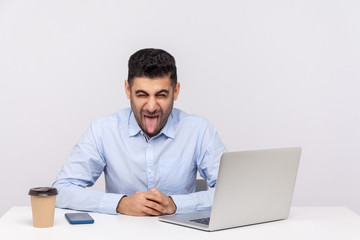 Crazy funny businessman sitting office workplace with laptop on desk, showing his tongue, looking at camera with naughty disobedient expression. indoor studio shot isolated on white background