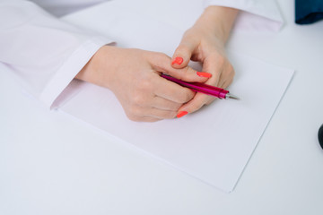 Close-up of hands of unrecognizable woman doctor holding pen under white empty paper. Hand of female doctor with pen writing on prescription blank