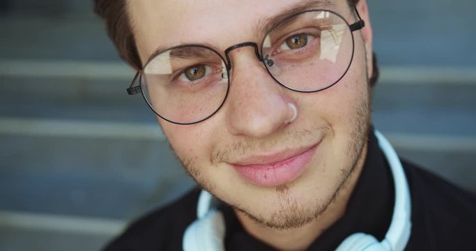 Close up portrait of joyful young guy in glasses with headphones on his neck smiling to camera outdoor. Caucasian handsome man on street with smile on face and deep look. Leisure concept