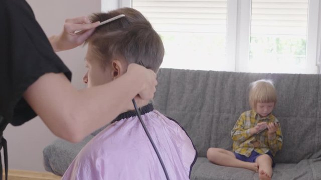 slender woman hairdresser cuts female. Blond boy on background sits on gray couch and play with smartphone
