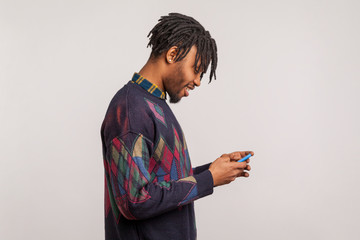 Profile portrait of happy trendy african guy with dreadlocks in sweatshirt using smartphone and smiling, pleased satisfied with connection quality. Indoor studio shot isolated on gray background