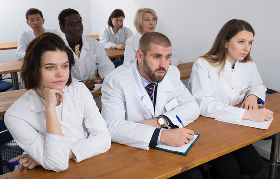Multinational group of people in white coats attentively listening and making notes while sitting in boardroom