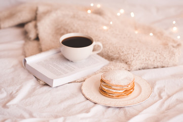 Cup of coffee with pancakes and open book on knitted sweater in bed. Warm and cozy atmosphere. Winter season. Good morning.
