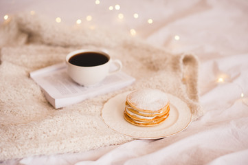 Cup of coffee with pancakes and open book on knitted sweater in bed. Warm and cozy atmosphere. Autumn season. Good morning.