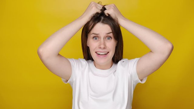 Portrait of hysterical young woman expression with her hands on head, stressed looking at camera, dressed in white t-shirt, isolated on yellow studio background. People emotion concept
