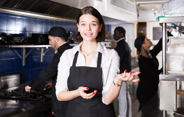 Cheerful young waitress showing modern kitchen of restaurant with working staff