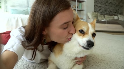happy smiling tender woman stroking, hugging and kissing corgi dog on selfie video Avki. girl cuddling puppy pet indoors. affectionate, love, enjoyment concept. pet owner look at phone camera